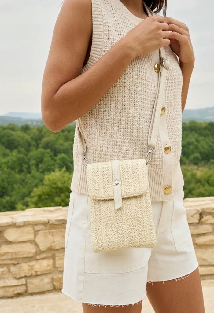 Person wearing a beige sleeveless top with gold buttons and holding a matching woven bag, standing against a scenic background.