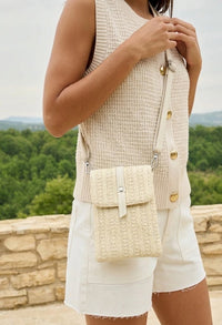 Person wearing a beige sleeveless top with gold buttons and holding a matching woven bag, standing against a scenic background.