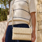 Woman wearing a cream sweater with navy stripes and holding a woven beige handbag.