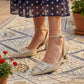 Beige high-heeled shoes with embellishments worn by a person in a navy polka dot dress on a patterned floor.