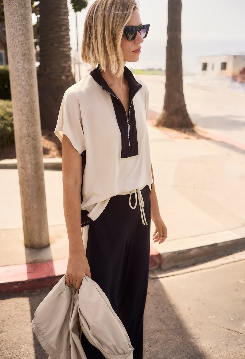 Woman wearing a white and black outfit with sunglasses, standing on a sidewalk.