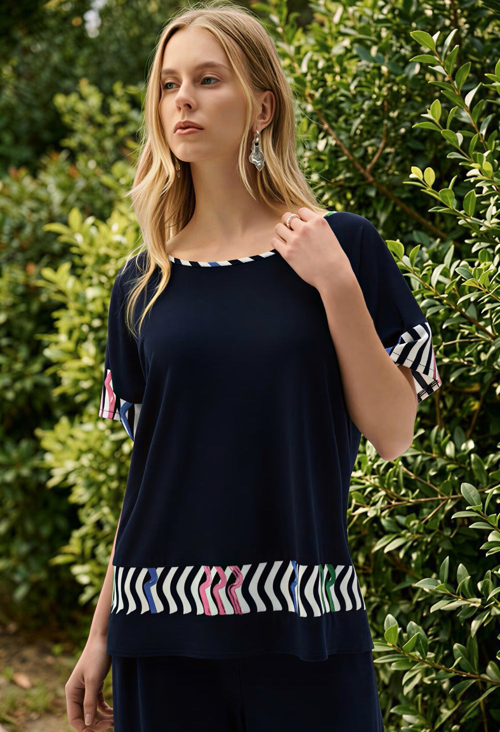 Woman wearing a navy blue top with colorful patterns in front of green foliage