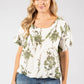 Woman wearing a white blouse with green leaf pattern and denim shorts on a gray background