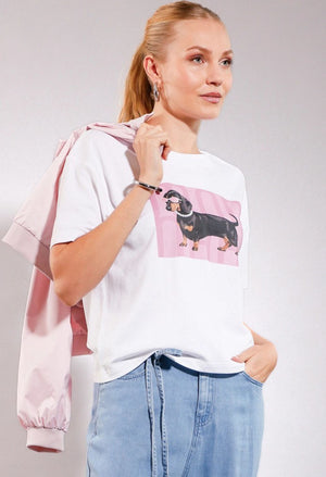 Woman wearing a white t-shirt with a dachshund graphic and a pink jacket on a gray background