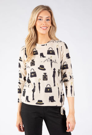 A woman modeling a cream-colored long-sleeve knit sweater with a black fashion-themed print of handbags, umbrellas, boots, and hats.