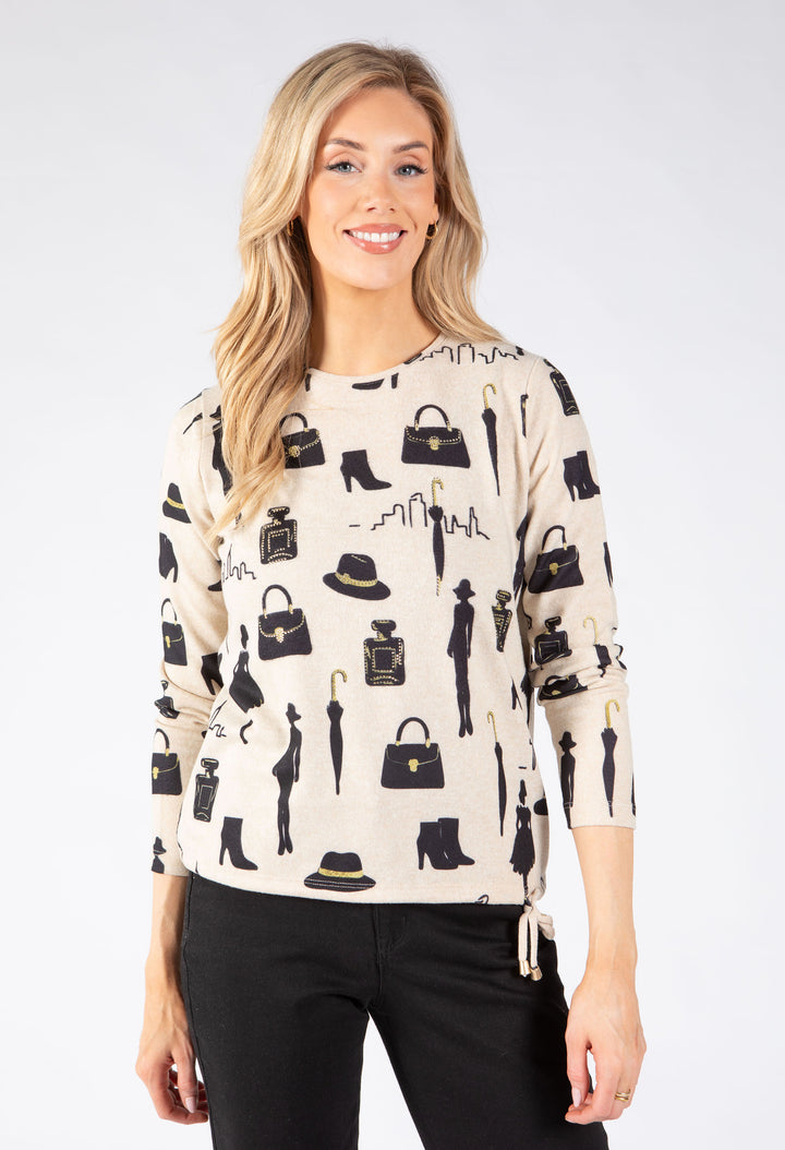 A woman modeling a cream-colored long-sleeve knit sweater with a black fashion-themed print of handbags, umbrellas, boots, and hats.