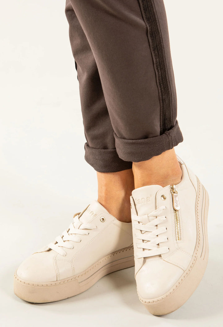A close-up of a person wearing off-white leather platform sneakers with side zippers and laces, paired with dark brown cuffed pants against a clean white background.