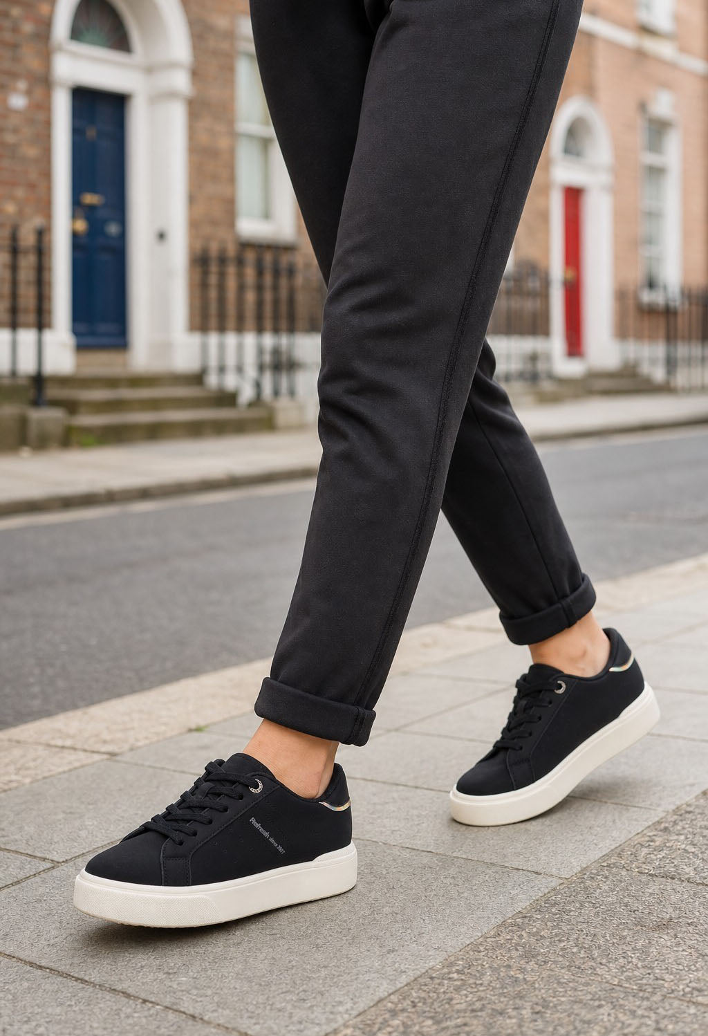 A person wearing black ankle-length trousers and black low-top sneakers with white soles, walking on a city sidewalk with brick buildings in the background.