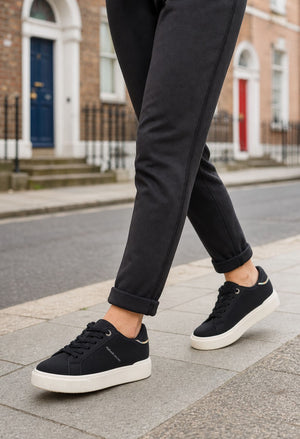 A person wearing black ankle-length trousers and black low-top sneakers with white soles, walking on a city sidewalk with brick buildings in the background.