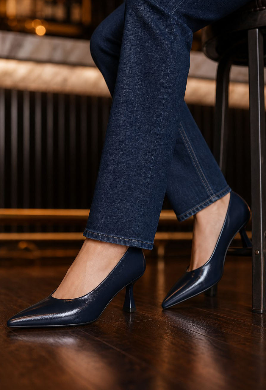 A person seated on a stool wearing dark navy pointed-toe high heels with dark blue jeans, photographed indoors on a wooden floor.