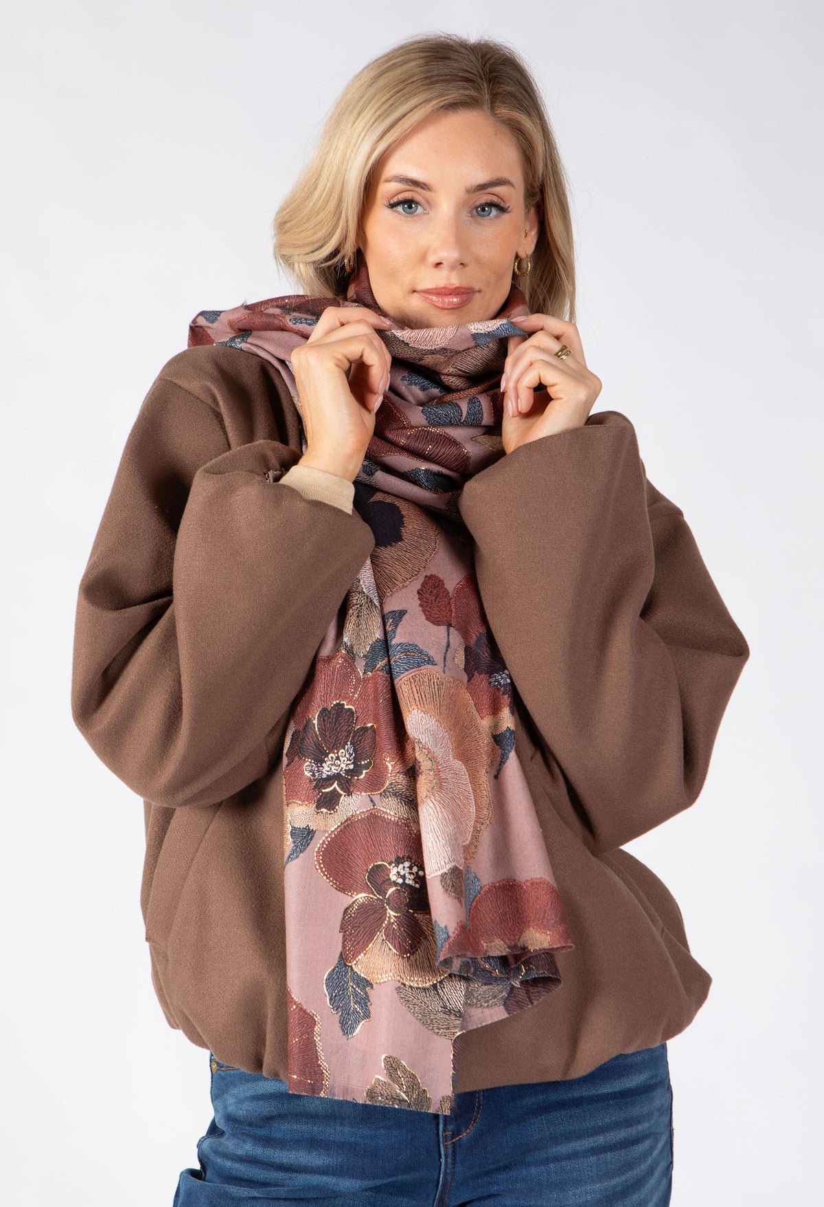 A woman wearing a brown jacket and blue jeans models a dusty pink floral scarf with brown, red, and blue tones against a light gray studio background.