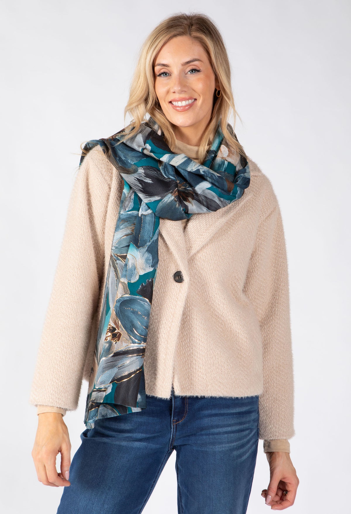 A woman modeling a beige textured cardigan and a blue and gray abstract print scarf, paired with blue jeans, against a plain white studio background.
