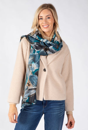 A woman modeling a beige textured cardigan and a blue and gray abstract print scarf, paired with blue jeans, against a plain white studio background.