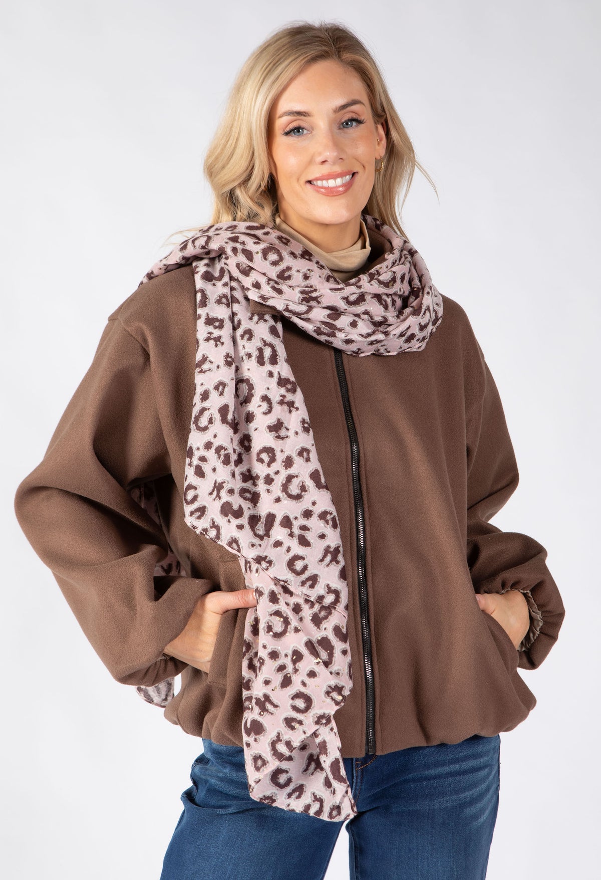 A woman modeling a brown zip-up jacket and a leopard-print scarf in front of a light gray studio background.
