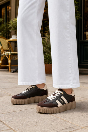 A person wearing white wide-leg jeans and dark brown platform sneakers with cream stripes, standing on a stone patio.
