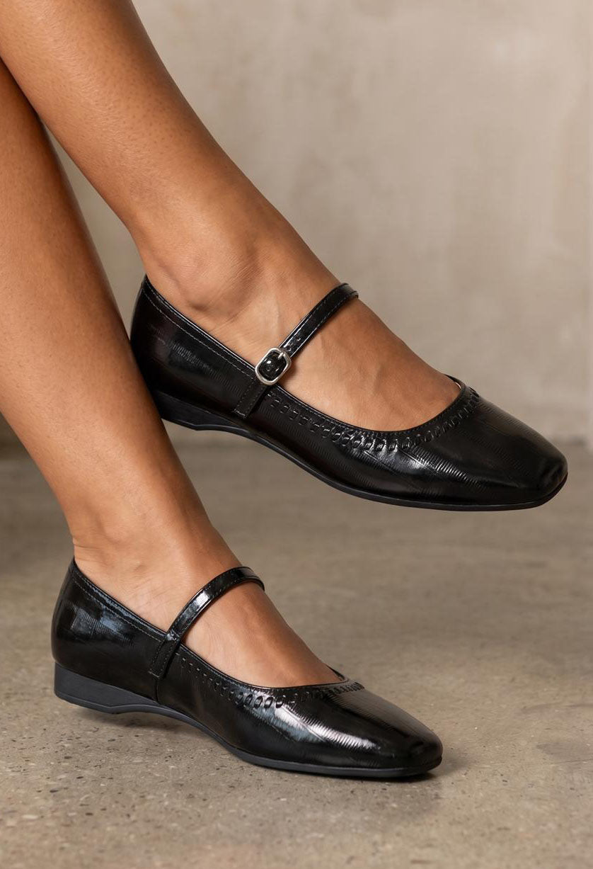 A pair of black patent leather Mary Jane flats with a square toe and a single strap with a silver buckle, shown on a model's feet against a neutral concrete background.