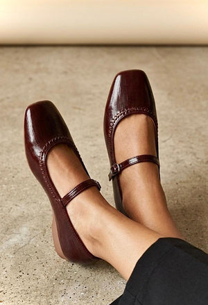 A pair of dark burgundy Mary Jane flats with a glossy finish, worn on a person's feet against a textured concrete floor.