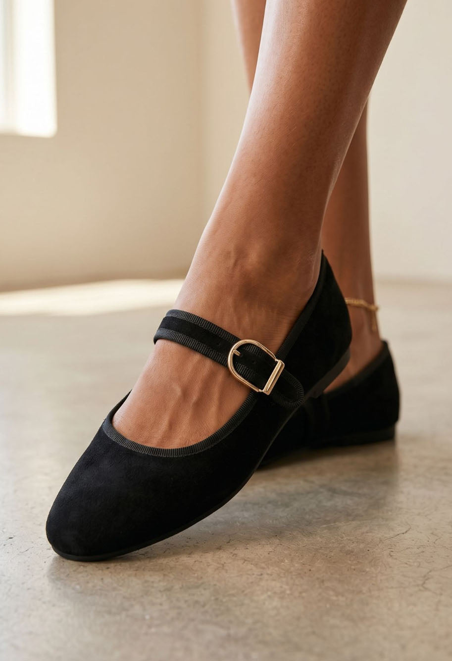 Black suede Mary Jane flats with a gold-tone buckle, shown on a model's feet in a softly lit indoor setting.