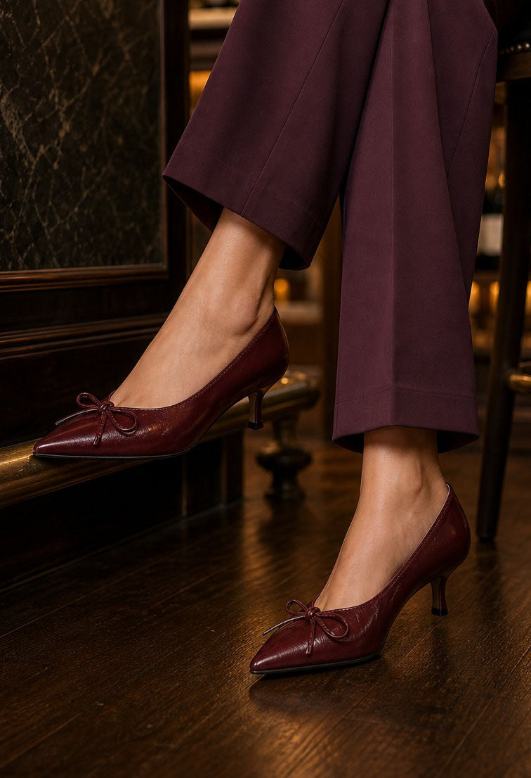 A close-up of a person wearing burgundy pointed-toe kitten heel pumps with small bow details, paired with dark purple wide-leg trousers, standing on a dark wood floor.