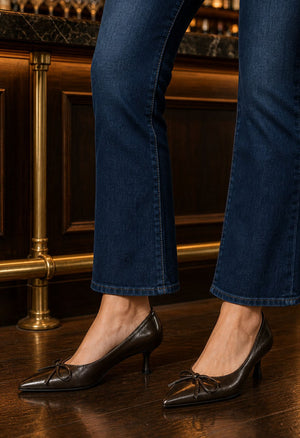 A person wearing dark brown pointed-toe kitten heel pumps with bow details, paired with dark blue jeans, standing on a polished wooden floor in front of a dark wood and brass bar.