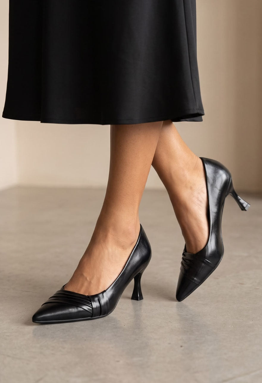 Black leather pointed-toe mid-heel pumps worn with a black midi skirt, shown on a model against a neutral beige background.