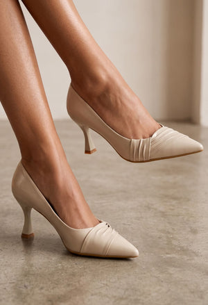 A pair of women's pointed-toe pumps in a light beige or cream shade, shown being worn on a model's feet against a neutral indoor background.
