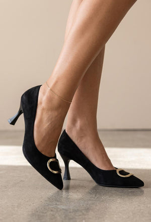 Black suede pointed-toe pumps with gold-tone oval hardware, shown on a model's feet against a neutral beige studio background.