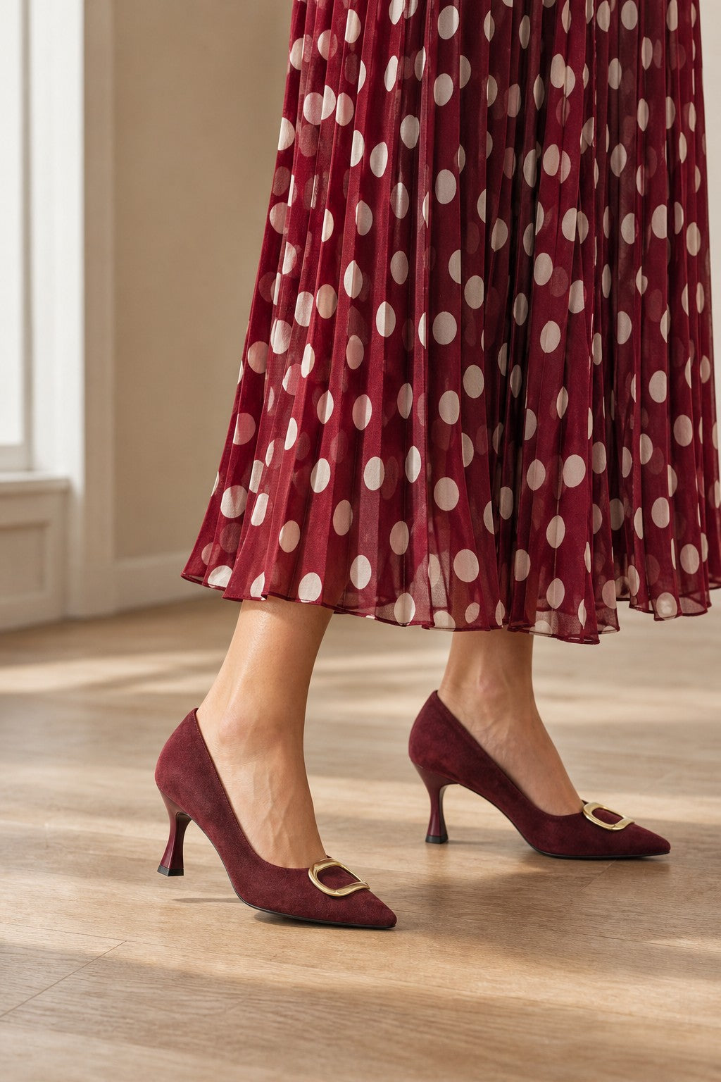 A person wearing burgundy pointed-toe kitten heel pumps with gold buckle details, paired with a burgundy pleated skirt featuring white polka dots, standing on a light wood floor.