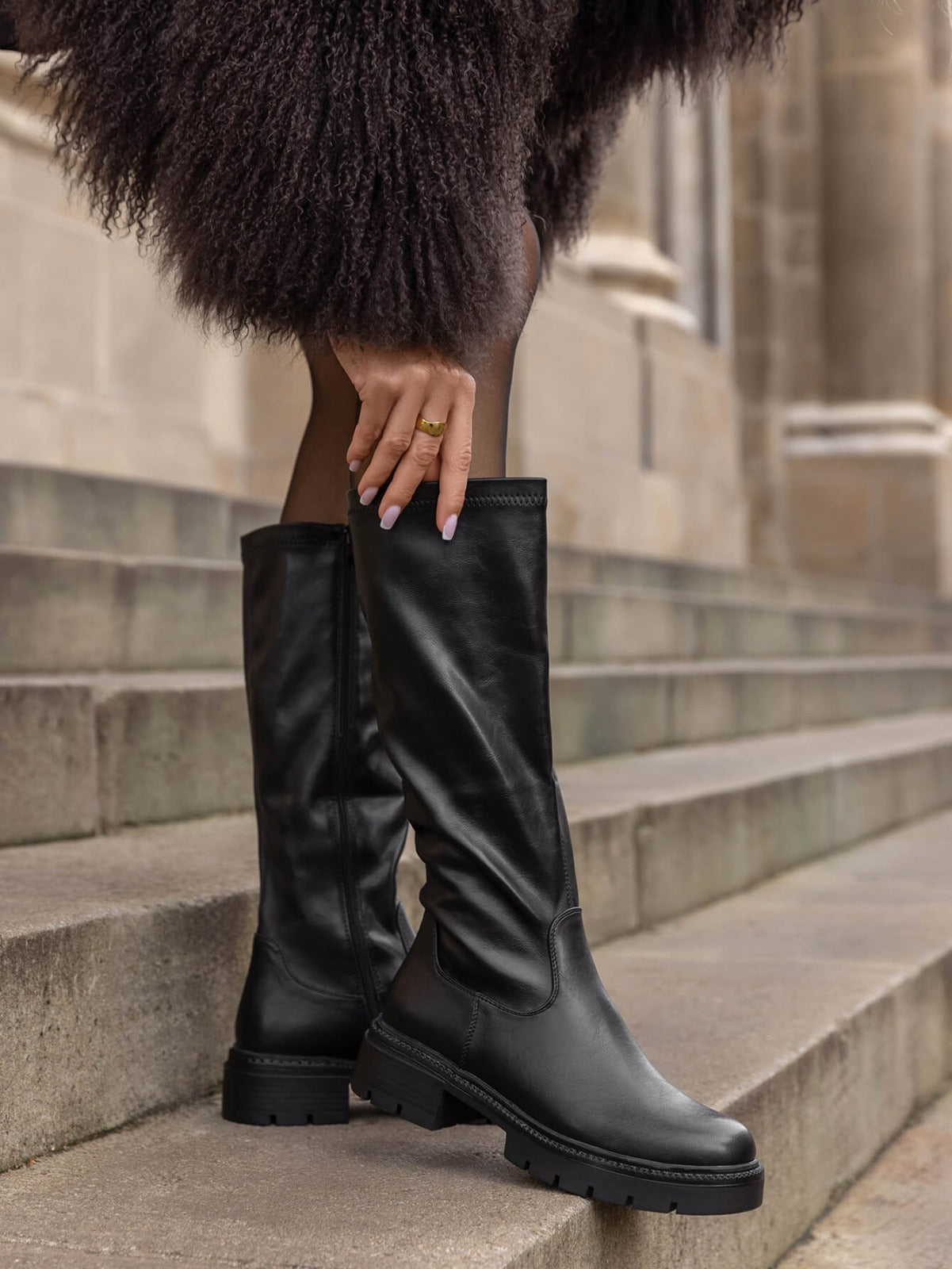 A person wearing tall black leather boots with a chunky lug sole, standing on stone steps. A hand with pink nails and gold rings rests on the hem of a dark brown textured coat.
