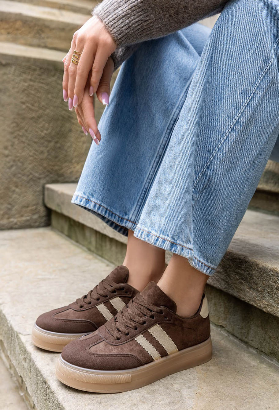 A person sitting on stone steps wearing brown suede sneakers with beige stripes, light blue jeans, and a gray knit sweater.