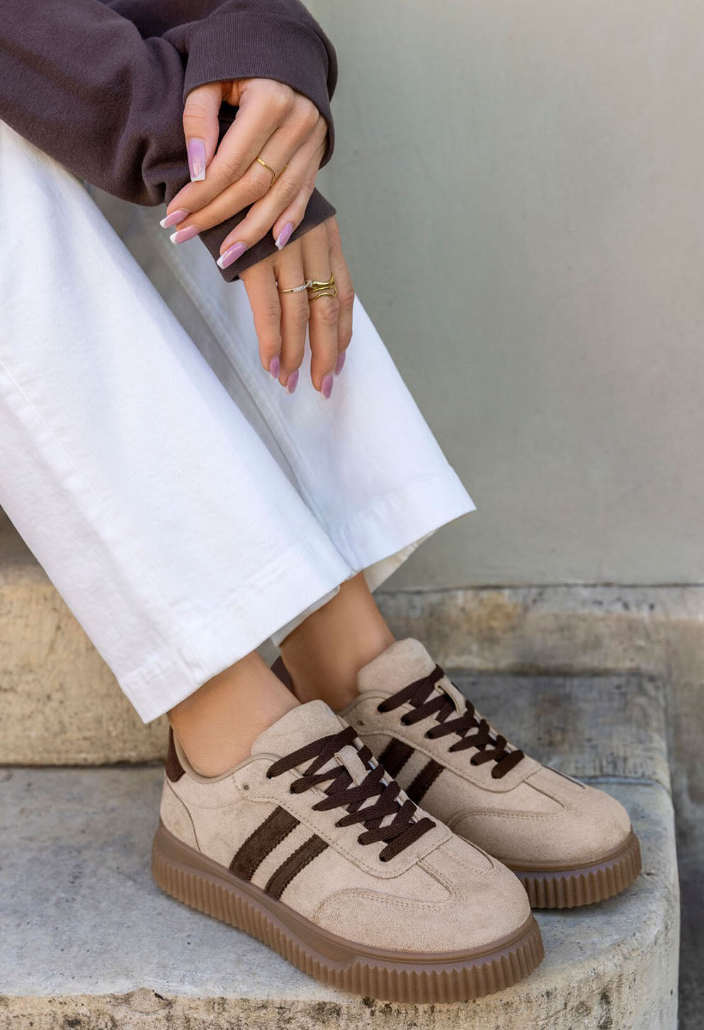 A person wearing beige low-top sneakers with dark brown laces and three dark brown stripes on the side, paired with white pants and a dark brown top, sitting on stone steps.