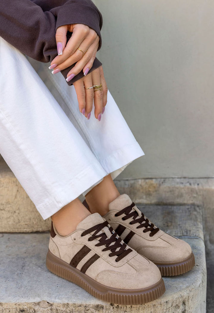 A person wearing beige low-top sneakers with dark brown laces and three dark brown stripes on the side, paired with white pants and a dark brown top, sitting on stone steps.