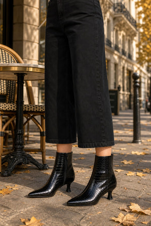 Black pointed-toe ankle boots with a glossy crocodile-embossed texture, worn with black cropped pants on a city sidewalk.