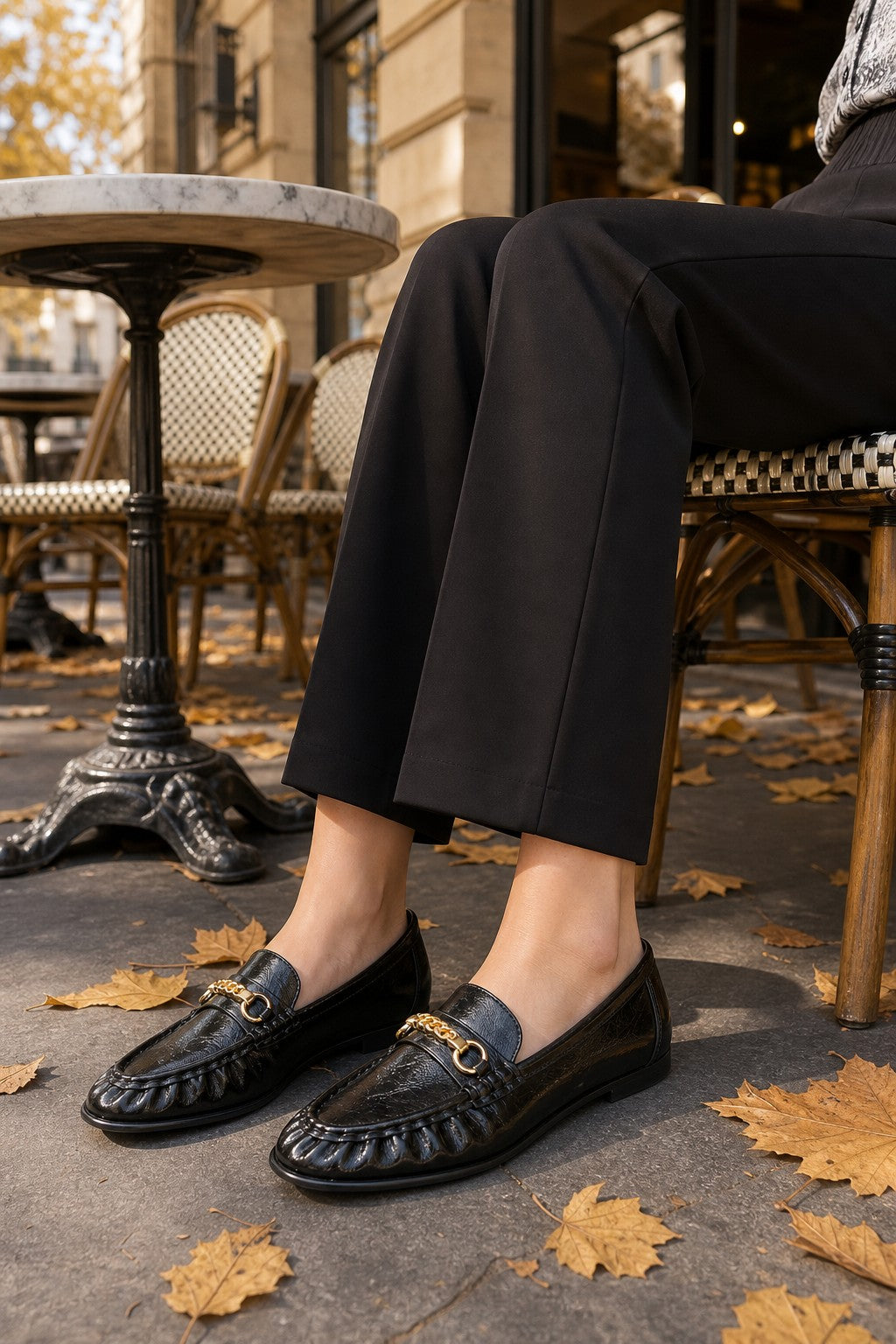 A person seated outdoors at a cafe wearing black leather loafers with gold-tone horsebit details, paired with black cropped trousers. Fallen autumn leaves are scattered on the pavement, and a black-and-white checkered chair is visible in the background.