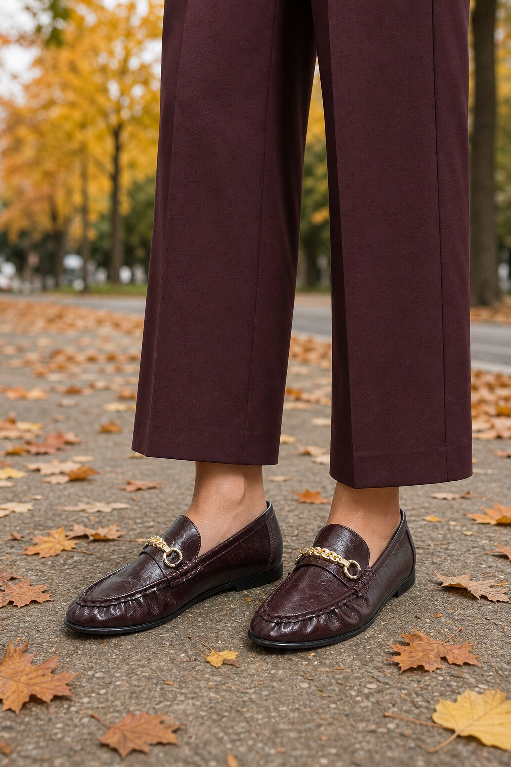 A person wearing dark burgundy cropped trousers and matching loafers with gold horsebit details, standing on a paved path scattered with autumn leaves outdoors.