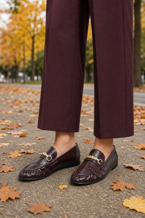A person wearing dark burgundy cropped trousers and matching loafers with gold horsebit details, standing on a paved path scattered with autumn leaves outdoors.