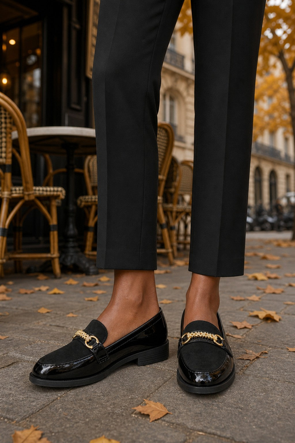 A person wearing black patent leather loafers with gold horsebit hardware, paired with black trousers, standing on a cobblestone street with cafe chairs and autumn leaves in the background.
