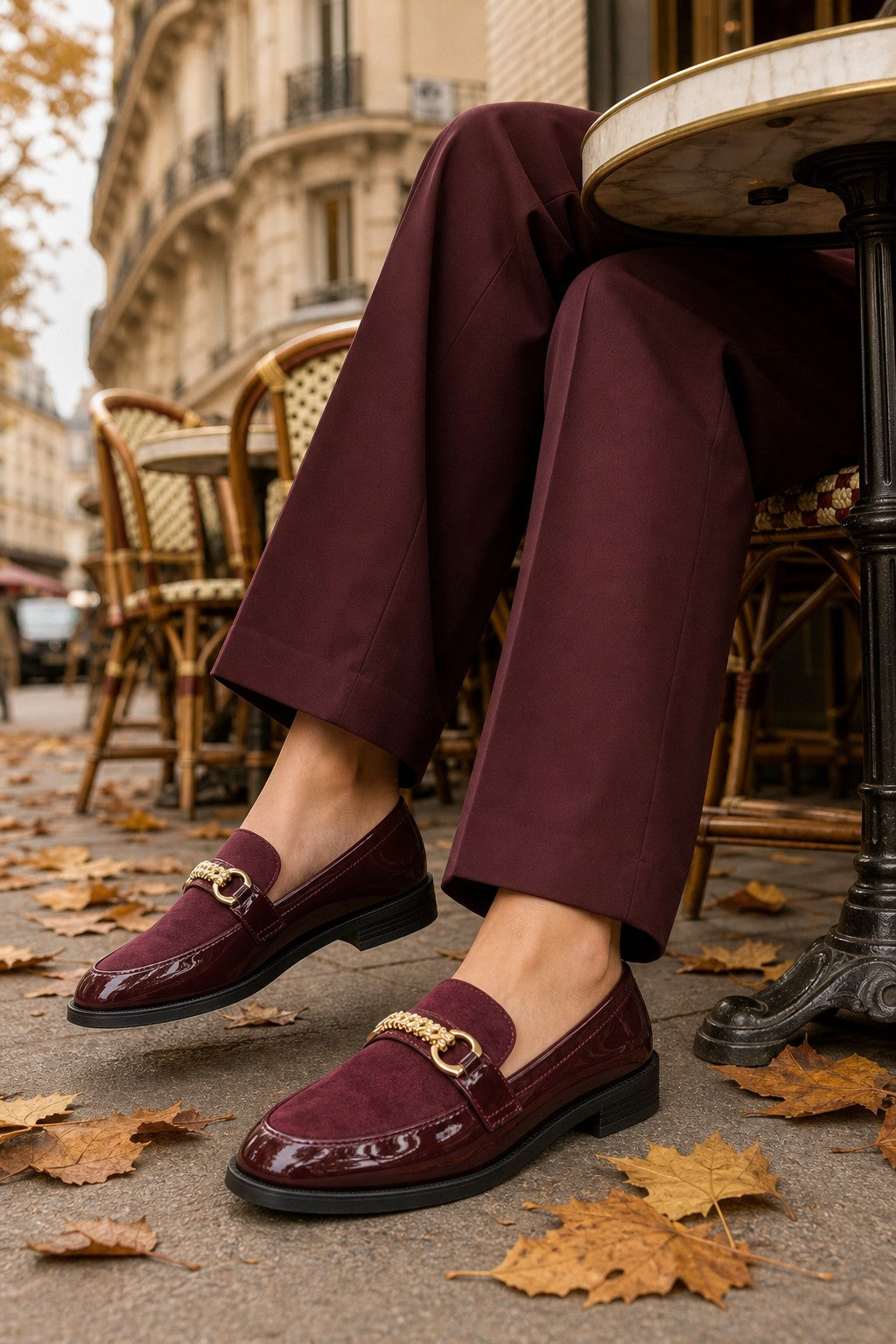 A person seated at an outdoor cafe table wearing burgundy loafers with gold-tone horsebit details and matching burgundy trousers, with fallen leaves on the ground and a Parisian-style building in the background.