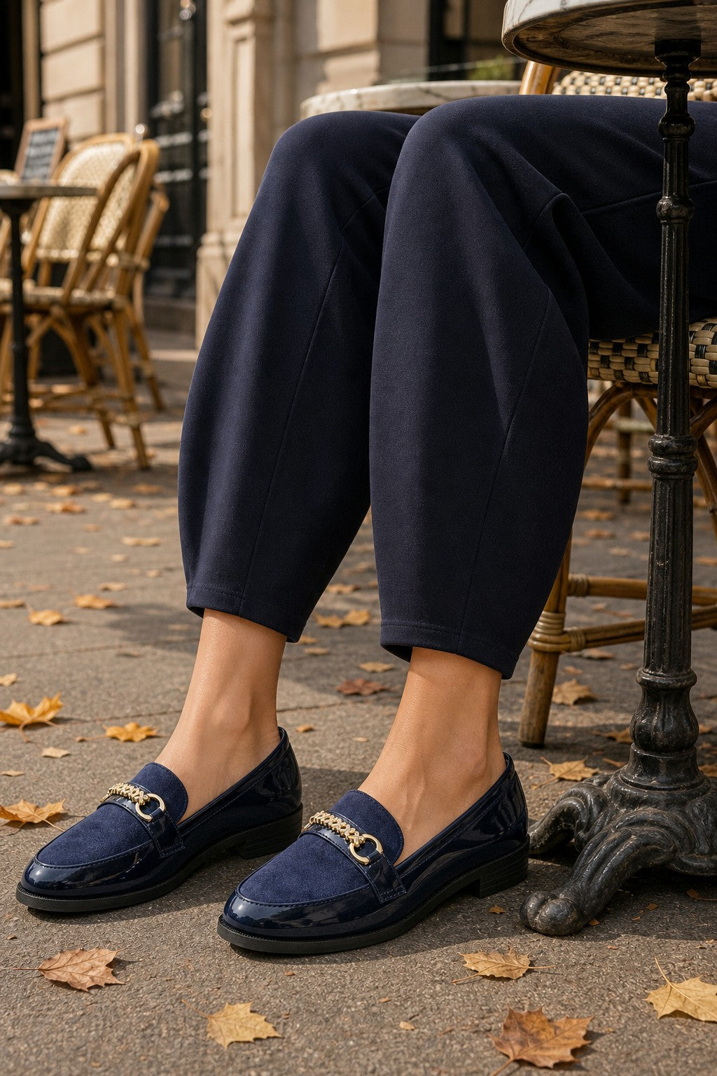 A person seated at an outdoor cafe table wearing navy loafers with gold horsebit hardware and dark navy pants, with fallen leaves on the pavement.