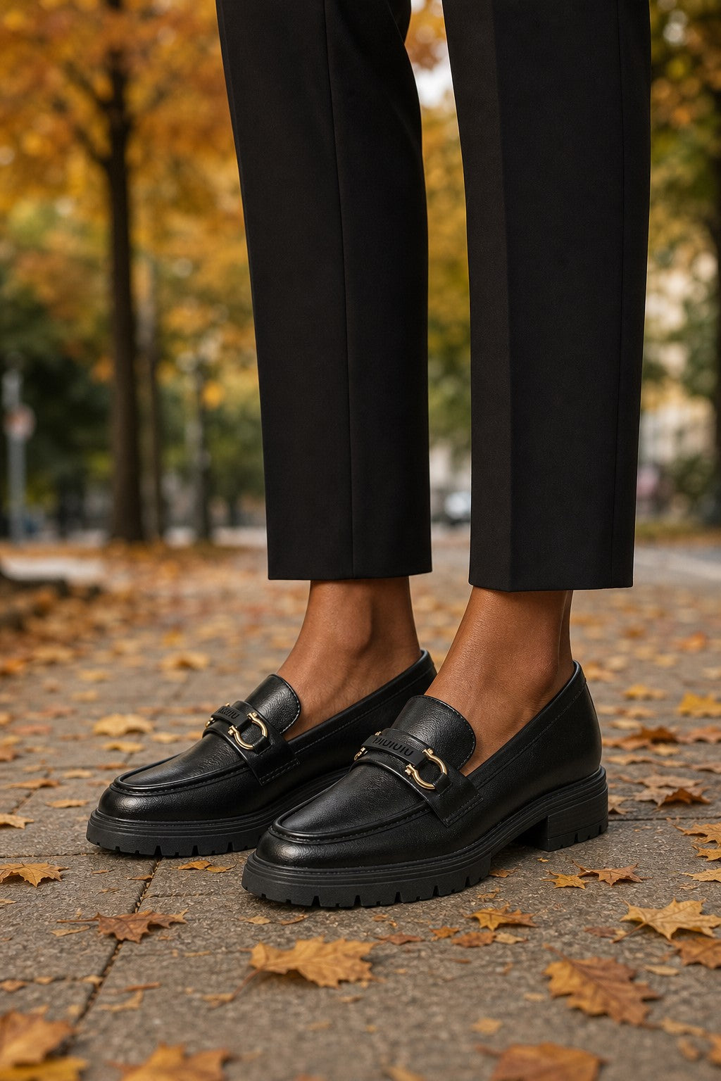 Black leather penny loafers with gold-tone buckle detailing, worn with black trousers on a paved path covered with fallen autumn leaves.