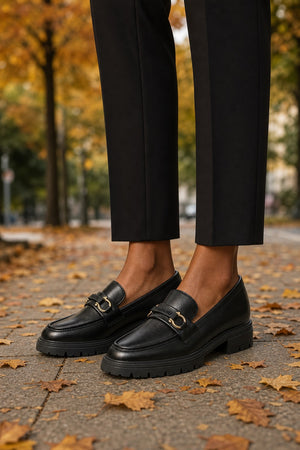 Black leather penny loafers with gold-tone buckle detailing, worn with black trousers on a paved path covered with fallen autumn leaves.