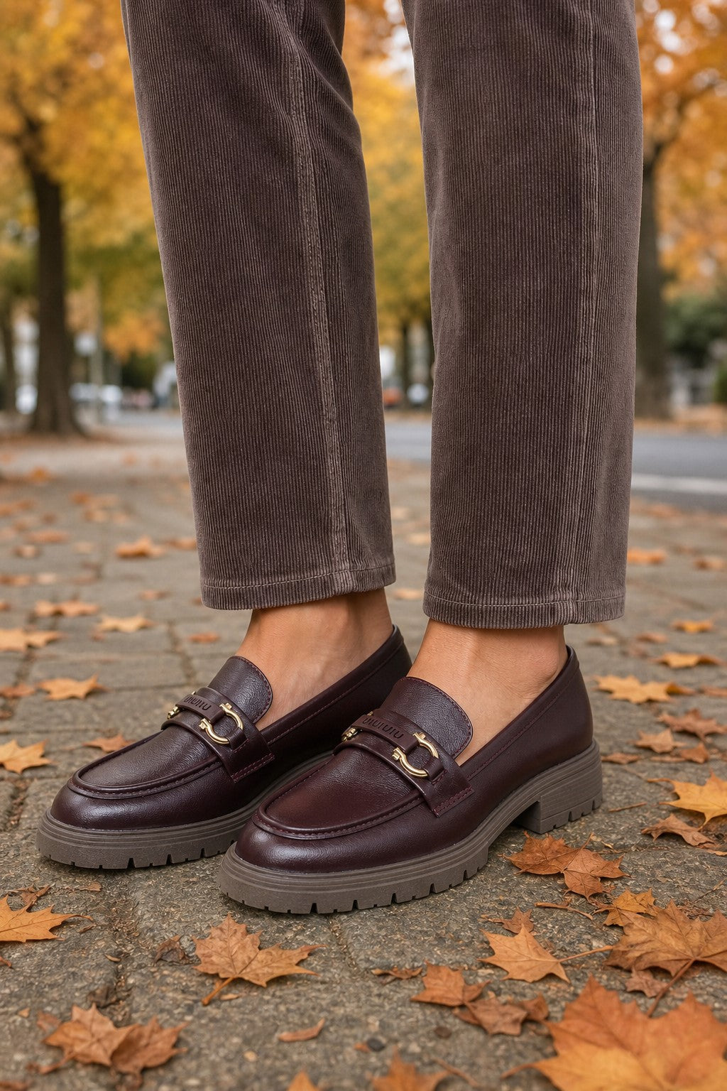 A person wearing dark brown corduroy trousers and dark burgundy leather penny loafers with gold-tone hardware, standing on a paved path scattered with autumn leaves.