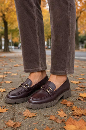 A person wearing dark brown corduroy trousers and dark burgundy leather penny loafers with gold-tone hardware, standing on a paved path scattered with autumn leaves.