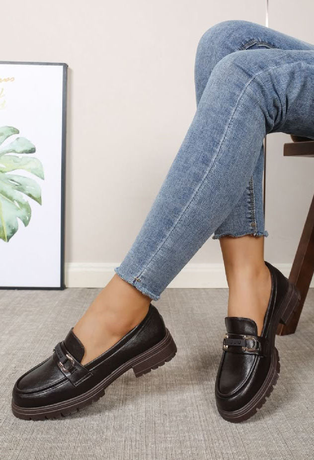 A person wearing black leather penny loafers with chunky brown soles and blue jeans, seated on a wooden chair in a room setting.