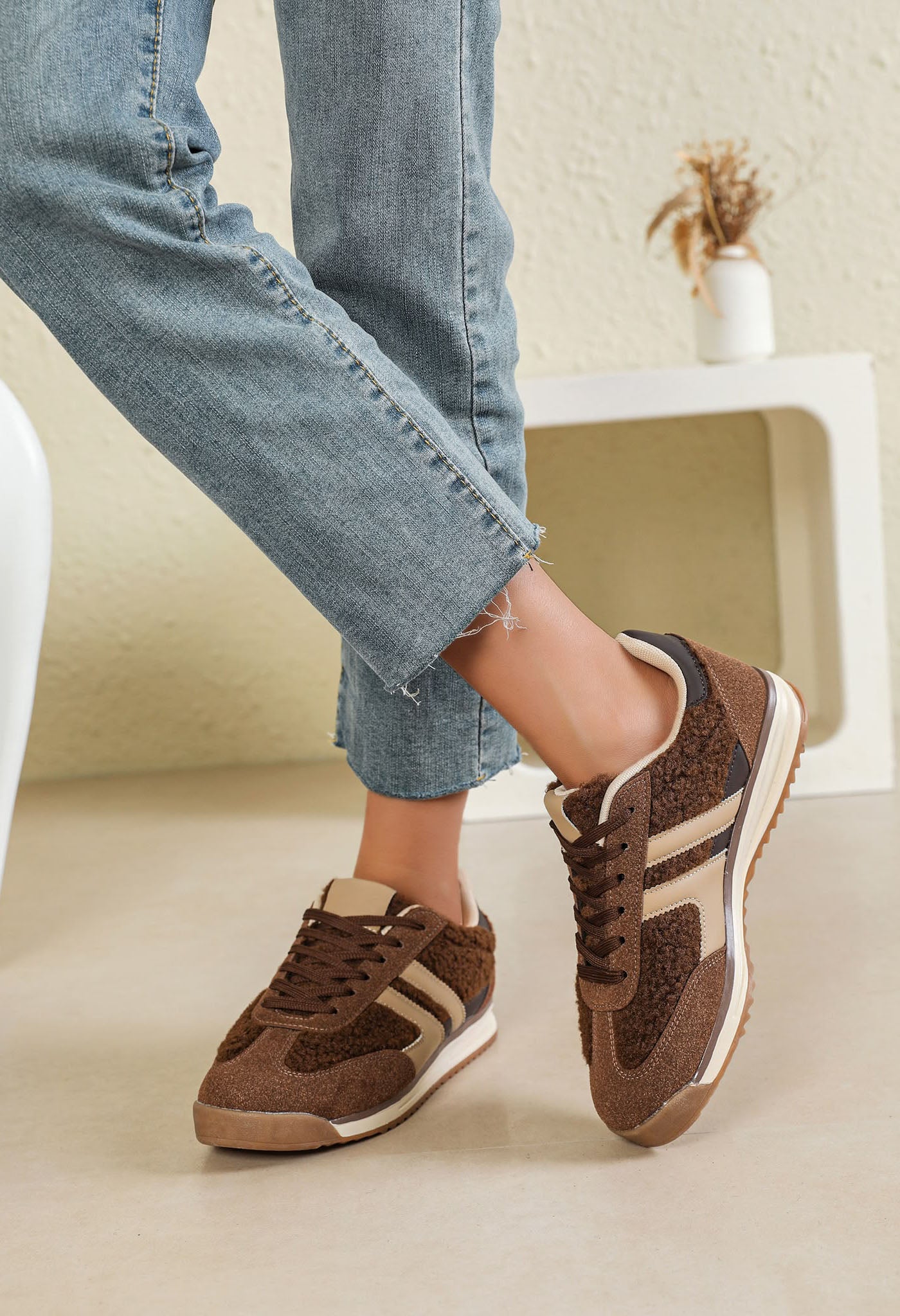 A person wearing brown and beige low-top sneakers with light blue jeans, standing in a minimalist indoor setting with a white shelf in the background.