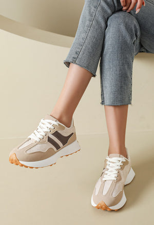 A person wearing beige and brown platform sneakers with white laces and white midsoles, paired with gray cropped jeans, seated against a warm beige background.
