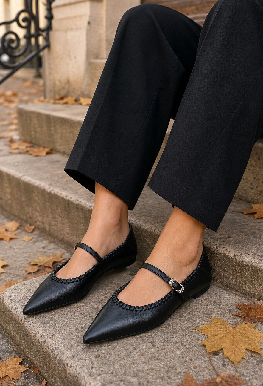 Black pointed-toe Mary Jane flats with braided trim and a single strap, worn with black pants on outdoor stone steps with scattered autumn leaves.