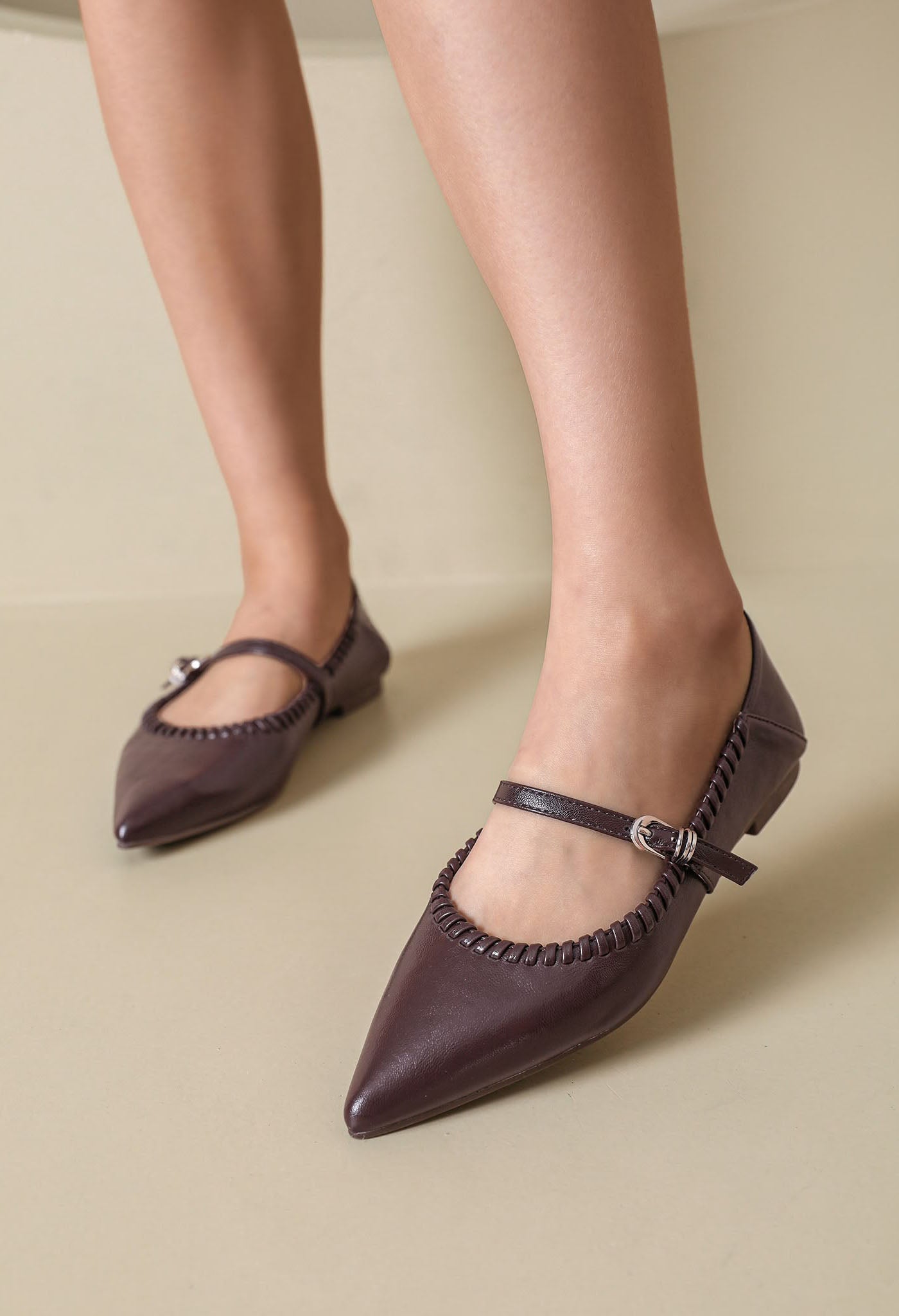 A pair of pointed-toe Mary Jane flats in a deep plum purple, worn on a model's feet against a neutral beige background.
