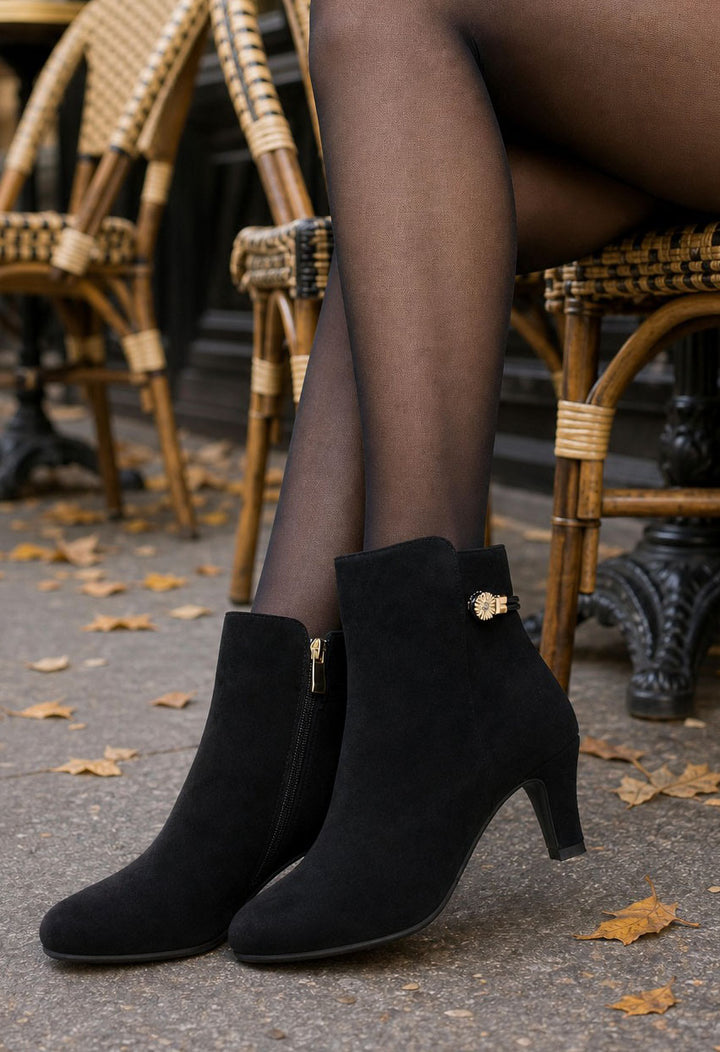 Black suede ankle boots with a small gold decorative accent on the side, worn with sheer black tights by a person seated on a wicker chair outdoors.