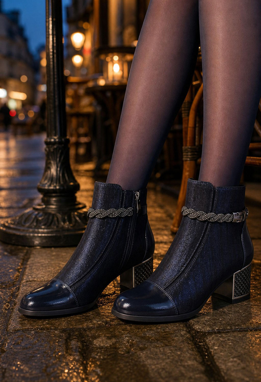 A close-up of a person's legs wearing dark navy blue ankle boots with a textured braided strap detail around the ankle, standing on a cobblestone street at night.